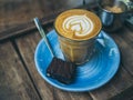 Piccolo Latte coffee topping with flower art from milk in small glass with a piece of homemade brownies cake on blue ceramic plate