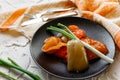 Pickled bell peppers of different colors, on a black matte plate, decorated with green onions, cutlery and a cloth napkin on