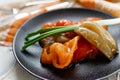 Pickled bell peppers of different colors, on a black matte plate, decorated with green onions, cutlery and a cloth napkin on