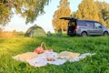 Picnic date in forest, tent and camp chairs, gray minibus with open door, blanket, wicker basket, wine glasses, snacks, fruit,