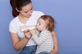 Picture of attarctive young mum feeding her little son from small bottle, mother holding kid in arms while posing isolated over Royalty Free Stock Photo