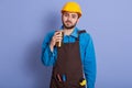 Picture of bearded handsome repairman with thermo mug in hand, wearing hard hat and special uniform, enjoying hot coffee or tea, Royalty Free Stock Photo