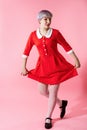 Picture of beautiful dollish girl with short light violet hair wearing red dress curtsey over pink background.