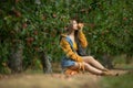 Picture of cheerful young lady sitting under apple tree holding and smells apple. Eyes closed