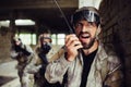 A picture of guy standing and leaning to wall. He is talking to a portable radio. His fighters are standing behind him