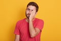 Picture of handsome man wearing casual red t shirt, standing with sad expression, covering half of his face with hand, looks tired Royalty Free Stock Photo