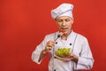 Picture of happy young senior chief cook in uniform standing isolated over red wall background, holding and eating salad