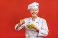 Picture of happy young senior chief cook in uniform standing isolated over red wall background, holding salad
