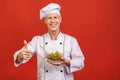 Picture of happy young senior chief cook in uniform standing isolated over red wall background, holding salad
