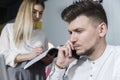 A picture of man looking forward and talking on the phone. He is serious. Girl is standing besides him and looking at journal. She