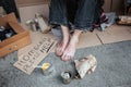 A picture of man`s feet without shoes. Man is sitting on cardboard. He is a beggar. There is a sign which says homeless