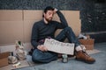 A picture of man sitting on the ground and holding homeless carboard in his hands. He is sad and tired. Guy is holding