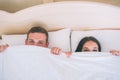 A picture of man and woman hiding faces under white blanket. They are lying together in one bed. Peple are looking up