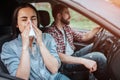 A picture of sick girl riding in car with young man. She is sneezing in napkin while he is paying attention to the road