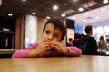 Picture of small girl sit alone at tabel and eat meal in cafe or restaurant. Hold burger or other food in hands and look Royalty Free Stock Photo