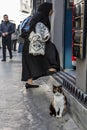 Stray cat standing in front of a shop while a muslim woman wearing the modest clothing with the traditional scarf is passing by