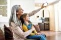 Picture time. Joyful grandmother smiling while taking selfie with her adorable little granddaughter, sitting on a couch