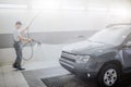 Picture of worker cleaning car. He uses water spray fo that. Young man holds flexible hose. He is serious and