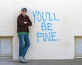 Seventy-three year old tourist posing by the words `You`ll Be Fine` on an outside wall in downtown Marfa, Texas.
