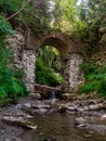 Old stone arched destroyed bridge over small river in a mountain forest
