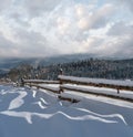 Picturesque waved shadows on snow from wood fence. Alpine mountain winter hamlet outskirts, snowy path, fir forest on far misty Royalty Free Stock Photo