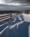 Picturesque waved shadows on snow from wood fence. Alpine mountain winter hamlet outskirts, snowy path, fir forest. High Royalty Free Stock Photo