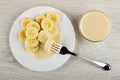 Banana strung on fork, white plate with slices of banana and condensed milk, bowl with milk on table. Top view