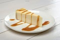 A piece of cheesecake, drizzled in caramel sauce on a white plate standing on wooden white table.