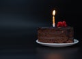Piece of chocolate birthday cake with one burning candle on white plate on deep black background. Birthday party Royalty Free Stock Photo
