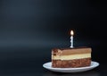 Piece of chocolate birthday cake with one burning candle on white plate on deep black background. Birthday party Royalty Free Stock Photo