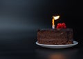 Piece of chocolate birthday cake with one burning candle on white plate on deep black background. Birthday party Royalty Free Stock Photo