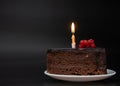 Piece of chocolate birthday cake with one burning candle on white plate on deep black background. Birthday party Royalty Free Stock Photo