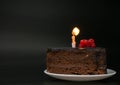 Piece of chocolate birthday cake with one burning candle on white plate on deep black background. Birthday party Royalty Free Stock Photo