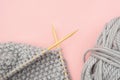 Piece of grey knitted fabric on bamboo wood needles with ball of yarn, process of knitting on pink background. Copy space for text