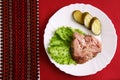 Piece of meat with slices of salted cucumber on white plate on the background of a red tablecloth with embroidered napkin