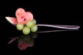 A piece of melon in the shape of a heart, fresh grapes and a piece of fig are placed on a metal spoon. The fruit salad is