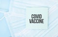 Piece of paper with phrase COVID VACCINE on pile of blue medical masks. Coronavirus pandemic concept