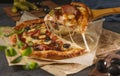 A piece of pizza with bacon, basil, olives, pickles and mozzarella cheese close-up on parchment paper on a dark background. Royalty Free Stock Photo