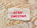 Piece of rough paper with an inscription in red pencil Merry Christmas lies on a smooth wooden board with an oak texture, strong Royalty Free Stock Photo