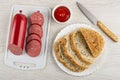 Sausage in polyethylene pack, slices of sausage on cutting board, bowl with ketchup, knife, slices of bread in plate on table. Top Royalty Free Stock Photo