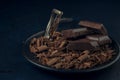 Pieces of chocolate, chocolate shavings and peeler in black plate on dark textured background