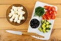 Pieces of cheese in saucer, bowls with dill, black olives, pieces of cucumbers, tomato, knife on cutting board on table. Top view Royalty Free Stock Photo