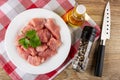 Pieces of raw pork meat in plate, bottle of vegetable oil on napkin, jar with condiment, knife on wooden table. Top view