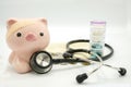 A piggy bank with a bandage at head and a stethoscope at chest with pill container and soft focus book bank on white background Royalty Free Stock Photo