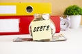 A glass jar with coins stands on a stack of dollar bills on the desktop. The jar has a sticker with the text DEBTS. Royalty Free Stock Photo