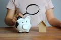 piggy bank and model wood house on table while woman using magnifying glass looking them , alternative save for home concept Royalty Free Stock Photo