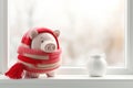 Piggy bank in red crochet scarf on white windowsill in winter. Heat saving, heating prices