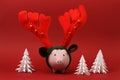 Piggy bank with reindeer horn and jingle bells standing on red background with three white origami trees Christmas background Royalty Free Stock Photo