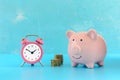 Piggy bank on a turquoise background, next to a small pink alarm clock and two stacks of coins. Royalty Free Stock Photo