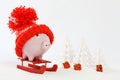 Piggy box with red hat with pompom standing on red sled on snow and around are snowbound trees and three gifts with gold bow Royalty Free Stock Photo